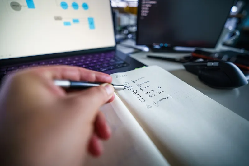 Hand writing a checklist next to a laptop displaying a workflow diagram