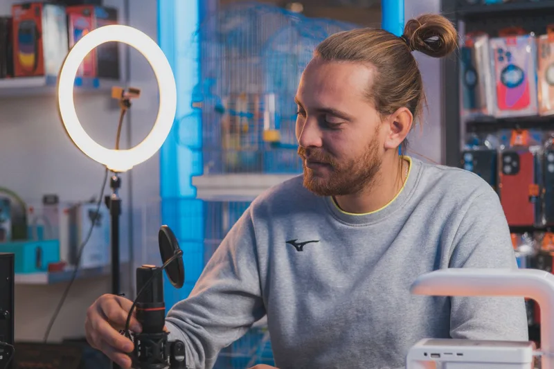 A man sitting in a studio setting adjusting a microphone with a ring light and equipment around